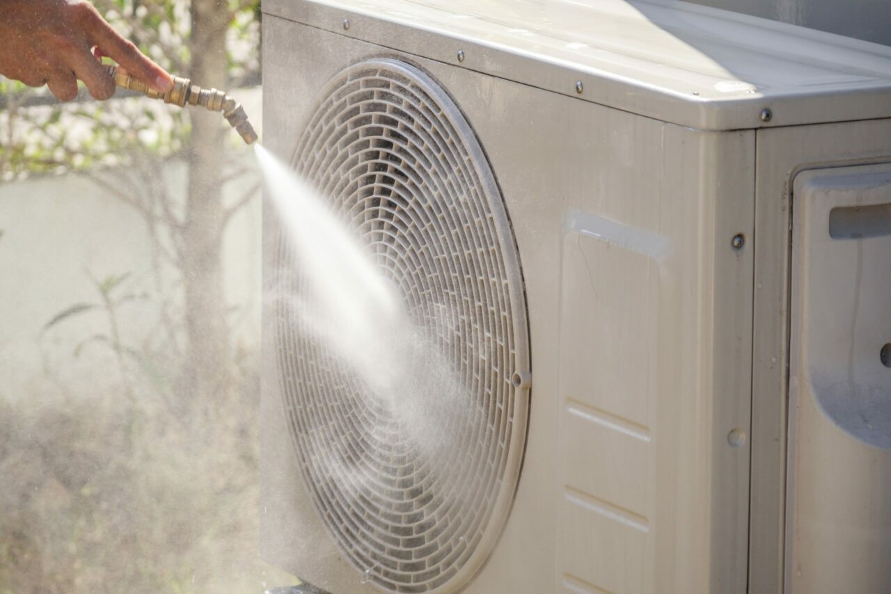 Person rinsing an outdoor AC condenser unit with a garden hose during seasonal cleaning maintenance