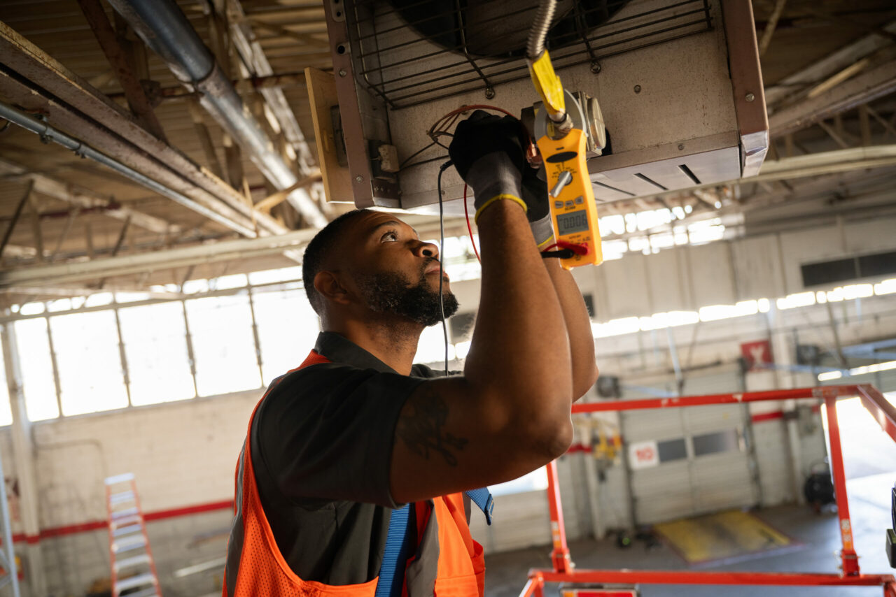HVAC technician using a clamp meter to test the wiring on a ceiling-mounted unit heater in a warehouse.