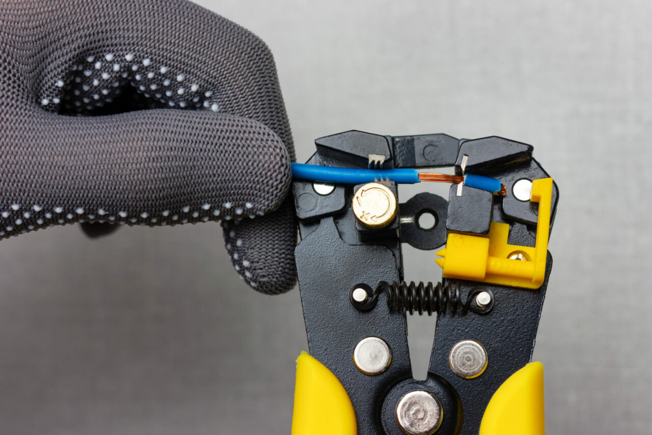An electrician wearing gray work gloves is stripping electrical wires with a stripper.