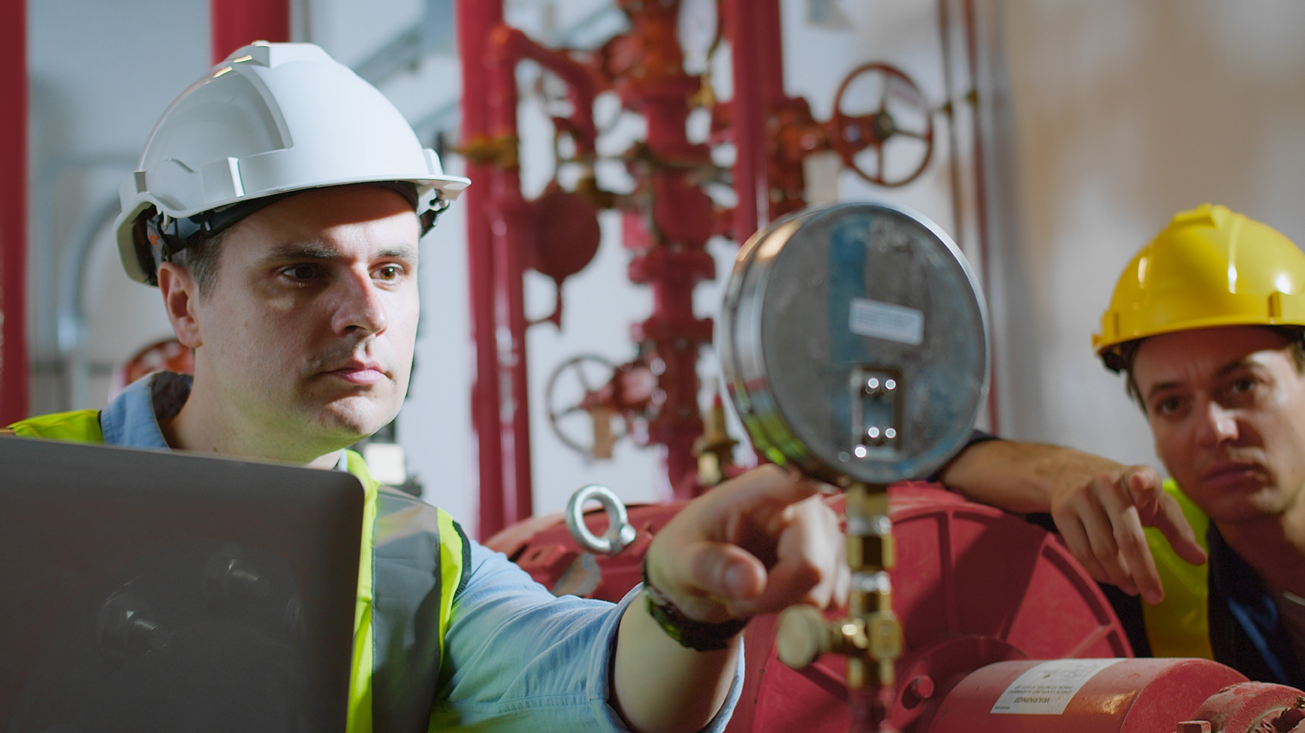 Industrial workers wearing hard hats inspect pressure gauges and equipment in a mechanical room during a safety check.