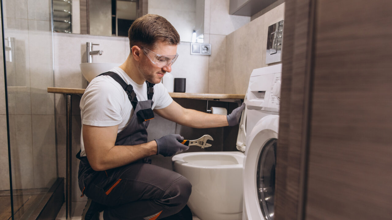 Person wearing safety goggles and gloves kneels beside a toilet, using a wrench to repair plumbing next to a washing machine in a bathroom.