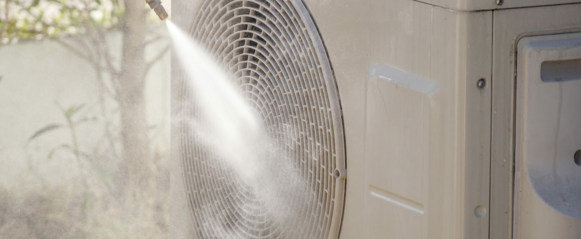 Person rinsing an outdoor AC condenser unit with a garden hose during seasonal cleaning maintenance