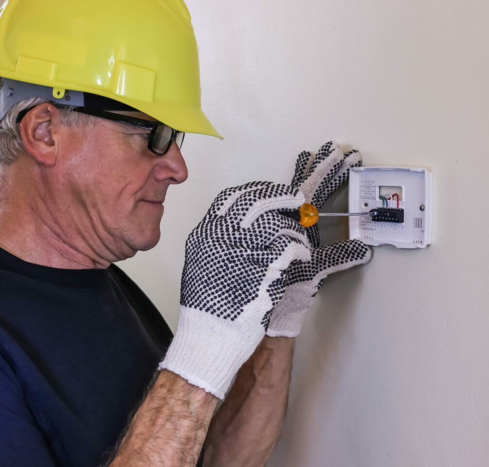 Thermostat base installation performed by HVAC technician wearing yellow hard hat and gloves.