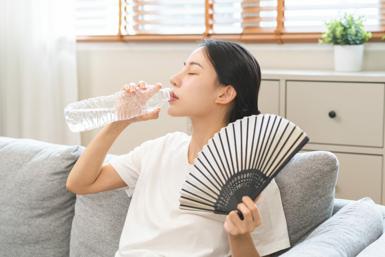 A woman sits on a couch drinking water, fanning herself to stay cool in a hot room