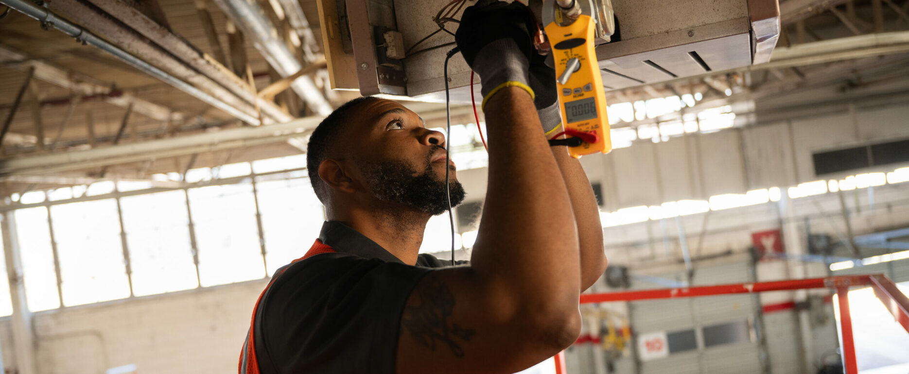 HVAC technician using a clamp meter to test the wiring on a ceiling-mounted unit heater in a warehouse.
