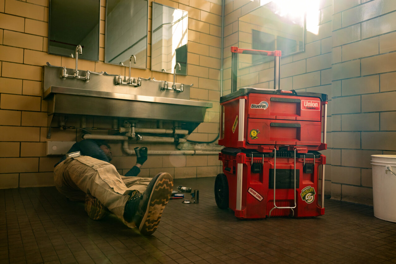 Professional plumber repairing plumbing under a sink in a tiled restroom with a red rolling toolbox to their right.