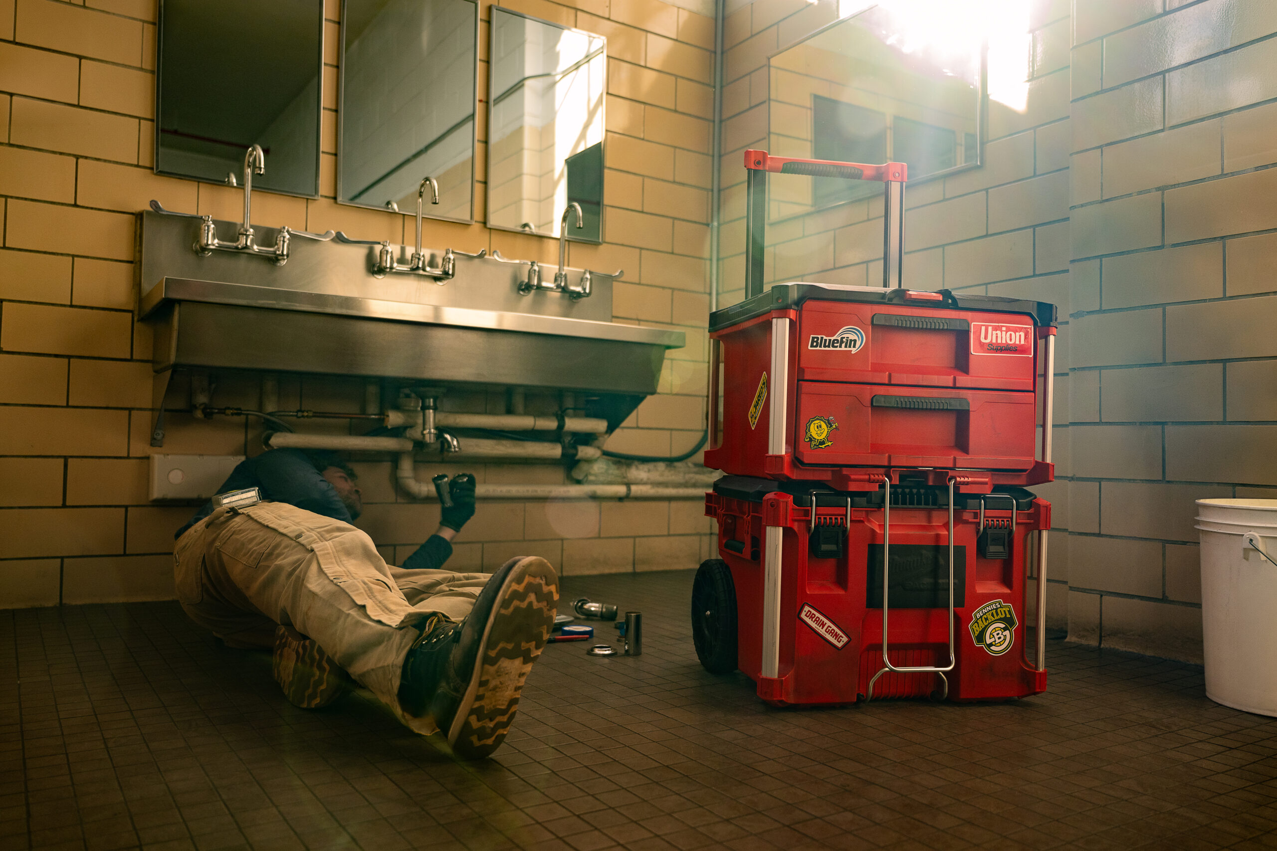 Professional plumber repairing plumbing under a sink in a tiled restroom with a red rolling toolbox to their right.